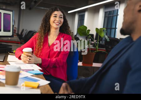 Happy diverse male and female colleagues having coffee and talking at desk in office Stock Photo