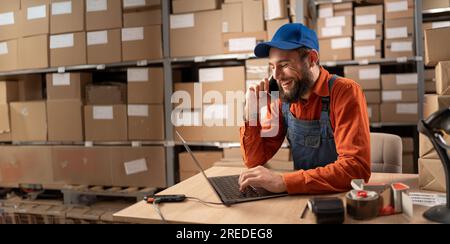 Warehouse worker doing inventory using laptop calling customers on mobile phone making a deal Stock Photo