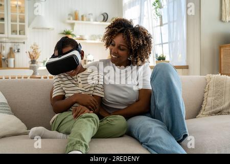 Happy joyful African American family mother and son using VR headset while relaxing together on sofa Stock Photo