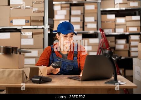 Delivery business. A young woman in a warehouse working with a laptop uses a barcode scanner enters data into the program Stock Photo
