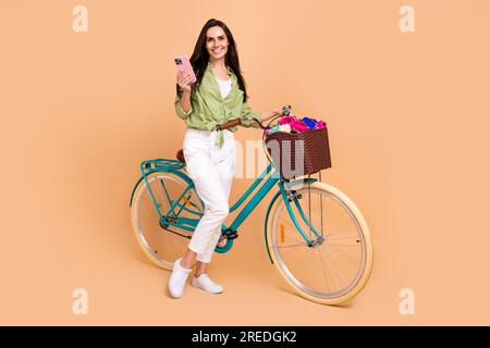 Full length photo of cute pretty woman wear green shirt white pants hold smartphone bicycle handlebars isolated on beige color background Stock Photo