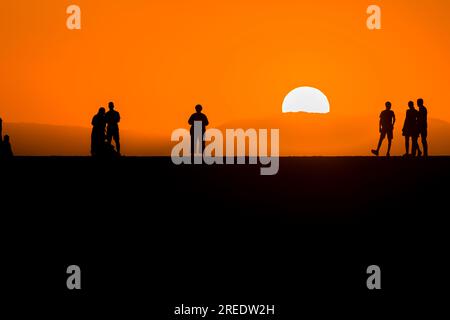 Silhouettes of people watching the sun set behind a distance mountain from a sea wall (Chania, Crete, Greece) Stock Photo