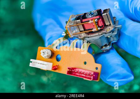 Closeup of optical pickup head with lens, coil springs and connectors on flex cables. Hand in blue glove holding read-write laser assembly from CD-DVD. Stock Photo