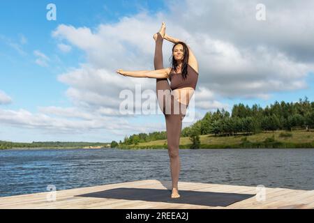 Woman practicing yoga doing variation of Urdhva Prasarita Eka Padasana exercise, vertical splits, balance asana, training in sportswear on the shore o Stock Photo