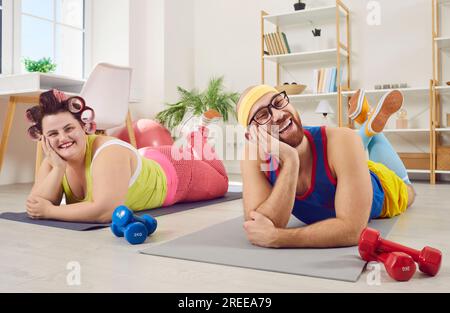 Funny couple in bright sportswear doing sport exercises at home lying on yoga mat with dumbbells. Stock Photo