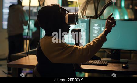 Cyber criminal using virtual reality haeadset to hack computer firewall, creating system malware and security breach to steal government information. Young spy doing hacktivism. Handheld shot. Stock Photo