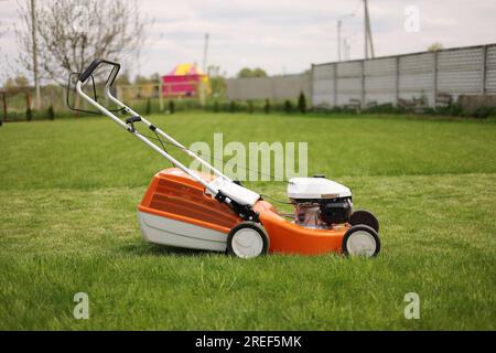 side view of modern orange-grey electric lawn mower cutting bright lush green grass. Gardening work tools. Rotary lawn mower machine on lawn. Professi Stock Photo