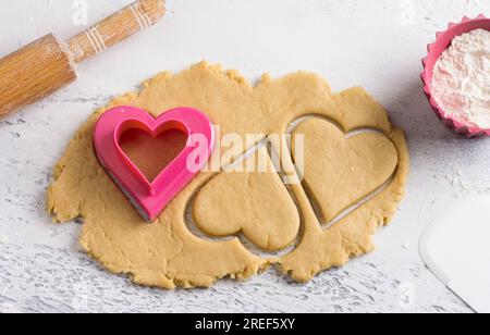 Cutted heart shaped dough cookies with cookie cutters, rolling pin and bowl of flour on a light blue tabletop, top view. Stage of cooking delicious ho Stock Photo