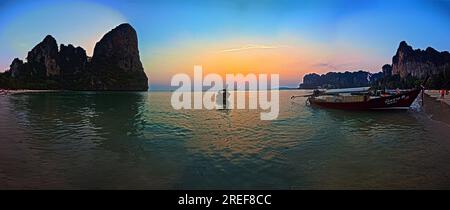 Panoramic shot of the sunset at Railay Beach in Krabi in cloudless sky framed by rocks and with typical longtail boat in the foreground photographed i Stock Photo