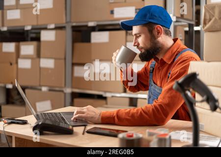 Professional male worker using laptop computer checking stock and inventory in the retail warehouse drinking hot coffee or tea Stock Photo
