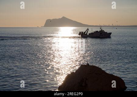Silhouettes in the Mediterranean of a fishing boat surrounded by seagulls and layers of the Calpe mountains Stock Photo