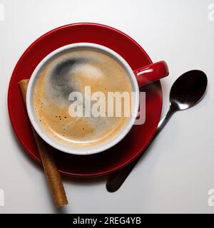 cup of coffee served in a mug on a red plate with a silver spoon and cinnamon stick Stock Photo