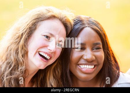 A heartwarming portrait capturing the strong bond between two female friends, one Caucasian and the other Black. Their heads are close together, and they share genuine smiles while looking away from the camera. This portrait showcases the beauty of friendship, unity, and shared happiness. Sisterly Bond: Smiling Portrait of Multi-Ethnic Female Friends. High quality photo Stock Photo