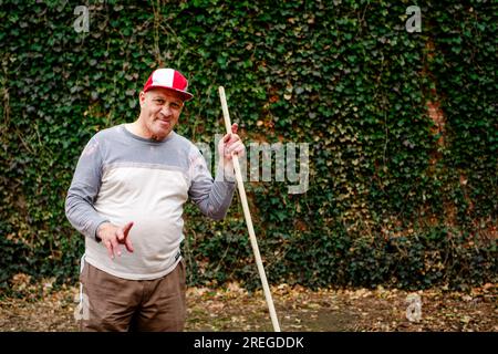 An smiling senior man stands with a rake in hand in front of ivy wall Stock Photo