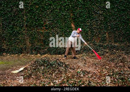 Side view of a senior man carrying a red rake in front of an ivy wall Stock Photo
