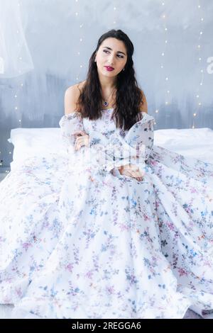 A girl on a bed in a white dress looks away on a gray background Stock Photo