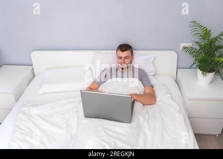 A man with laptop is lying in bed. Freelancer works remotely from home. Distance education. Online shopping without leaving home. Internet surfing. Gambling addiction. Black Friday sale. Cyber Monday. Stock Photo