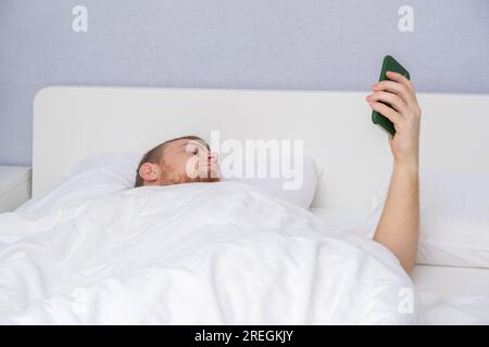 A man with a beard uses phone in his home bed. Shopping for goods without getting out of bed. The concept of people's dependence on gadgets. Internet addiction. Dependence on phone. Gadget addiction. Stock Photo