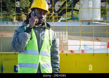 African ethnicity man with beard, disappointed construction worker talking on the phone, he is wearing yellow safety helmet and reflective vest, copy Stock Photo