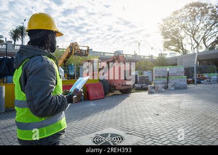 rear view of young black man of african ethnicity, construction worker supervising construction site materials, wears yellow safety helmet, reflective Stock Photo