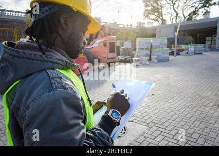 african ethnicity man engineer, wearing yellow safety helmet and reflective vest, checking construction site materials list taking note. copy space. Stock Photo