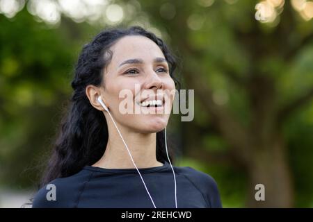 Young beautiful woman during active physical exercise in the park, rests and breathes fresh air, Hispanic woman in headphones uses an app on her phone to listen to music and online podcasts. Stock Photo