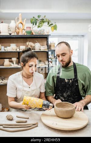 Couple of artisans in aprons drying clay bowl with heat gun on wooden board in ceramic workshop Stock Photo