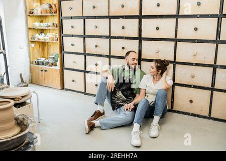 Cheerful couple of potters in aprons holding hands while sitting near cupboard in ceramic studio Stock Photo