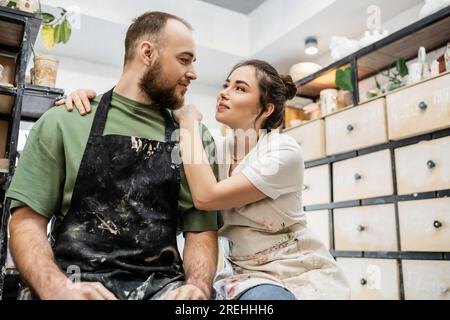 Craftswoman hugging and looking at boyfriend in apron while working in ceramic workshop Stock Photo