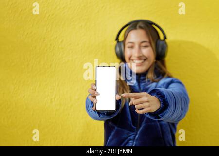 Latin girl with headphones out of focus shows the blank screen of her mobile phone isolated on a yellow background. Stock Photo