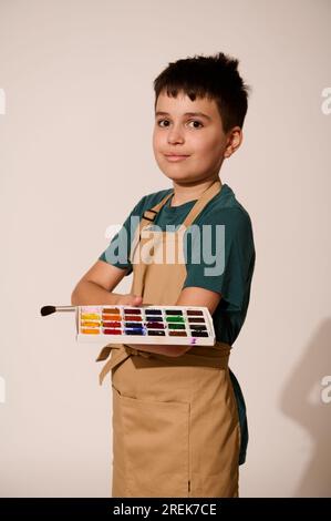 Vertical portrait of adorable teenage boy wearing beige apron, holding paintbrush and palette with watercolors, smiling, looking at camera, standing w Stock Photo