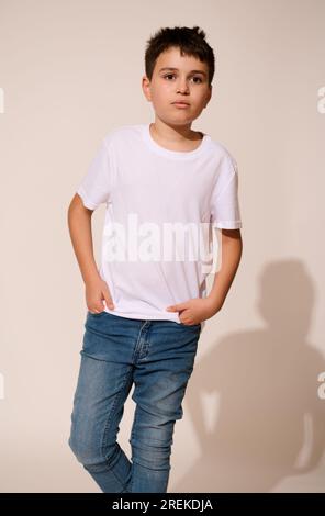 Authentic portrait of a Hispanic teenage boy in white t-shirt and blue jeans, putting his hands in pockets, thoughtfully looking aside, isolated on wh Stock Photo