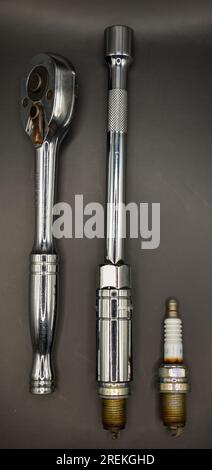 A socket wrench, an extension bar with a spark plug socket attached and two used spark plugs photographed against a dark background. Stock Photo