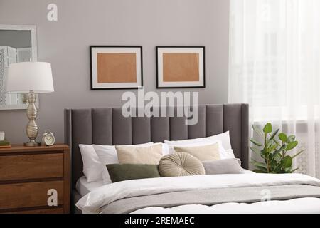 Comfortable bed with cushions, lamp and different decor on wooden chest of drawers in room. Stylish interior Stock Photo