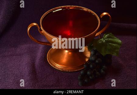 Golden colored chalice with Red liquid and grapes next to it on top of a purple cloth Stock Photo