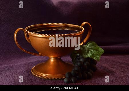 Golden colored chalice with Red liquid and grapes next to it on top of a purple cloth Stock Photo