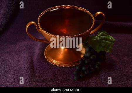 Golden colored chalice with Red liquid and grapes next to it on top of a purple cloth Stock Photo