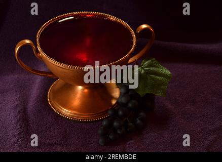 Golden colored chalice with Red liquid and grapes next to it on top of a purple cloth Stock Photo