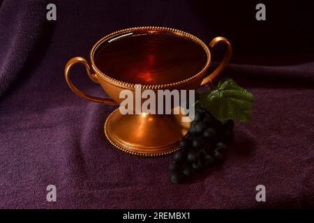 Golden colored chalice with Red liquid and grapes next to it on top of a purple cloth Stock Photo