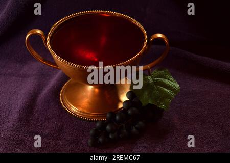 Golden colored chalice with Red liquid and grapes next to it on top of a purple cloth Stock Photo