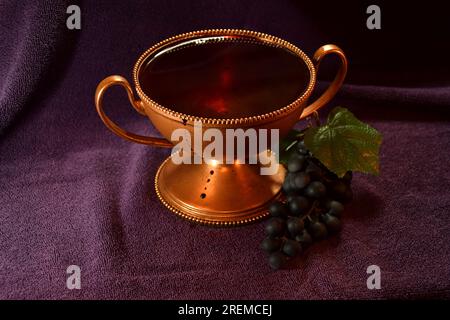 Golden colored chalice with Red liquid dripping down the side and grapes next to it on top of a purple cloth Stock Photo