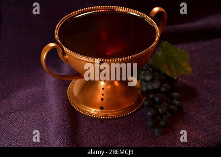 Golden colored chalice with Red liquid dripping down the side and grapes next to it on top of a purple cloth Stock Photo