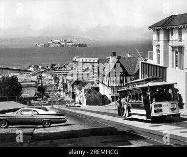San Francisco, California, November, 1965 The Powell and Hyde Street cable car as it climbs to the top of Russian Hill with Alcatraz Island in the background. Stock Photo