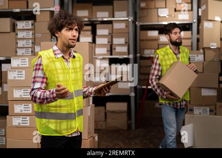 Team of two warehouse workers shipping order detail on digital tablet check goods on shelves with goods inventory in factory warehouse. Logistic Stock Photo