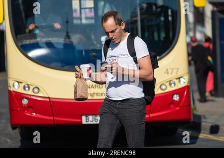 Man with rucksack or backpack, crossing road in front of bus, busy city, using mobile phone, with coffee and breakfast, in Summer, UK. Stock Photo