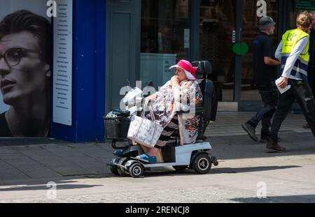 Large senior woman in electric mobility scooter on pavement at shops, carrying shopping bags, wearing summer clothes and hat in a city in England, UK. Stock Photo