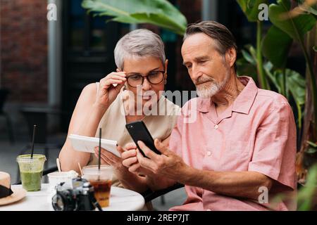 Aged woman looking at her husband's smartphone in an outdoor cafe. Senior man showing his wife something on his smartphone. Stock Photo