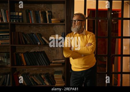 Pensive senior man standing with arms crossed on chest at home library Stock Photo