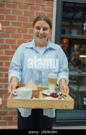 Happy smiling handsome woman waiter in a restaurant holding a tray with a latte coffee, cappuccino and berry cake in a restaurant Stock Photo