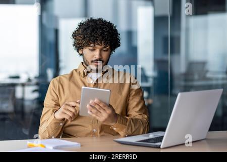 Serious focused businessman working with tablet computer sitting at desk inside office, hispanic thinking with laptop, man solving technical problem, using online application. Stock Photo